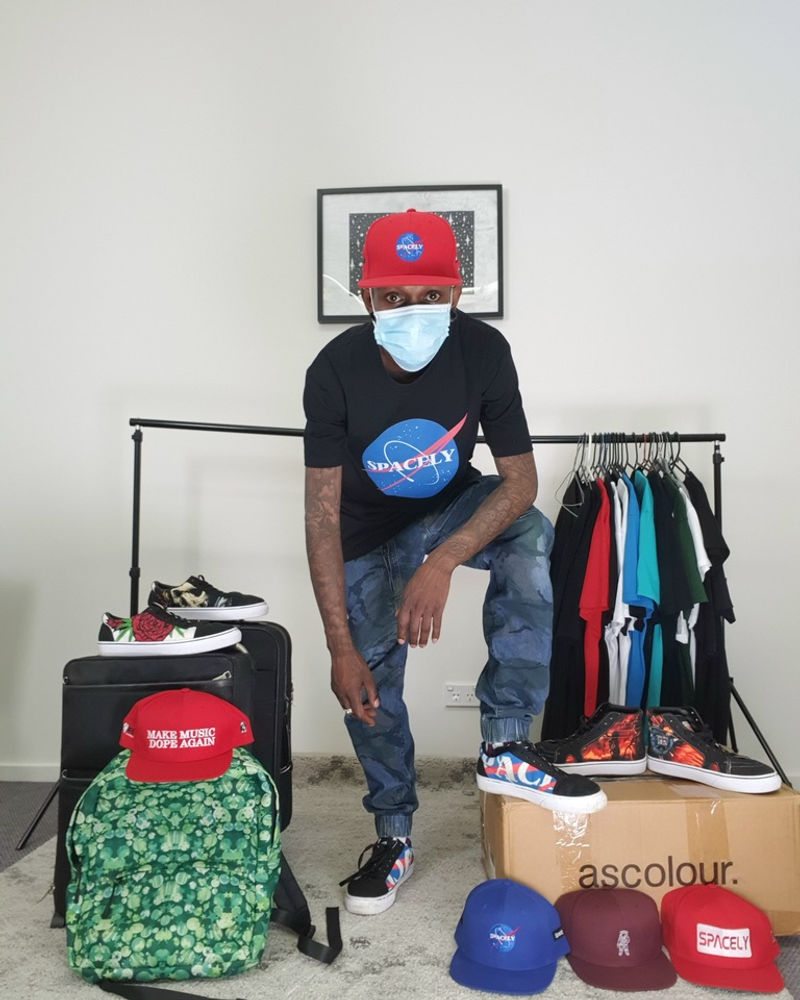 Man seated in a Spacely tee with a rack of clothing and caps behind him
